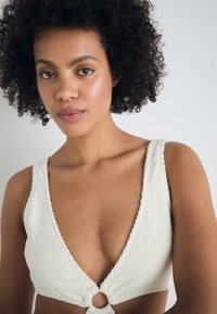 Bralette en tricot couleur crème avec un décolleté en V profond et de larges bretelles, mettant en avant un détail en nœud circulaire au centre. Tissu texturé.
