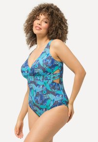 Maillot de bain une pièce bleu foncé avec des motifs de feuilles vertes et d'oiseaux. Présente des détails froncés et des découpes latérales. Fabriqué en matériau extensible.