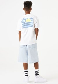 Camiseta de algodón blanca con un diseño gráfico de olas azules y un sol en la parte posterior. Combinada con pantalones cortos de mezclilla azul claro y zapatillas blancas.