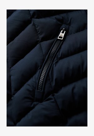 Marineblaue, gesteppten Jackenstoff mit einer Nahaufnahme eines seitlichen Reißverschlusses, der einen schlanken silbernen Ziehgriff und ein strukturiertes Polsterungsmuster zeigt.