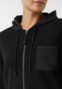Schwarze Zip-Hoodie mit einer Fronttasche, verstellbare Kapuze, glatter Textur und silbernem Reißverschluss-Detail.