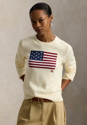 Kvinde iført en cremefarvet sweater med strikket amerikansk flag og initialerne "RL", kombineret med beige bukser og et flettebrunt bælte.