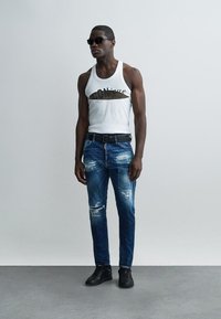 Uomo con occhiali da sole neri, maglietta bianca senza maniche con scritta "Iconique", jeans blu strappati, cintura nera e sneakers nere, in piedi davanti a uno sfondo semplice.