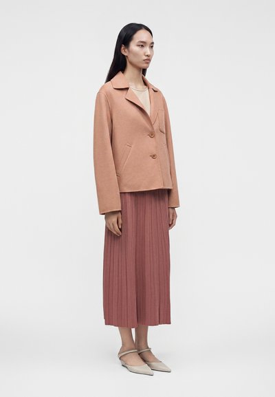 Femme aux longs cheveux noirs portant une veste marron clair, un top beige, une jupe midi plissée rose poudré et des ballerines beiges pointues avec des brides.