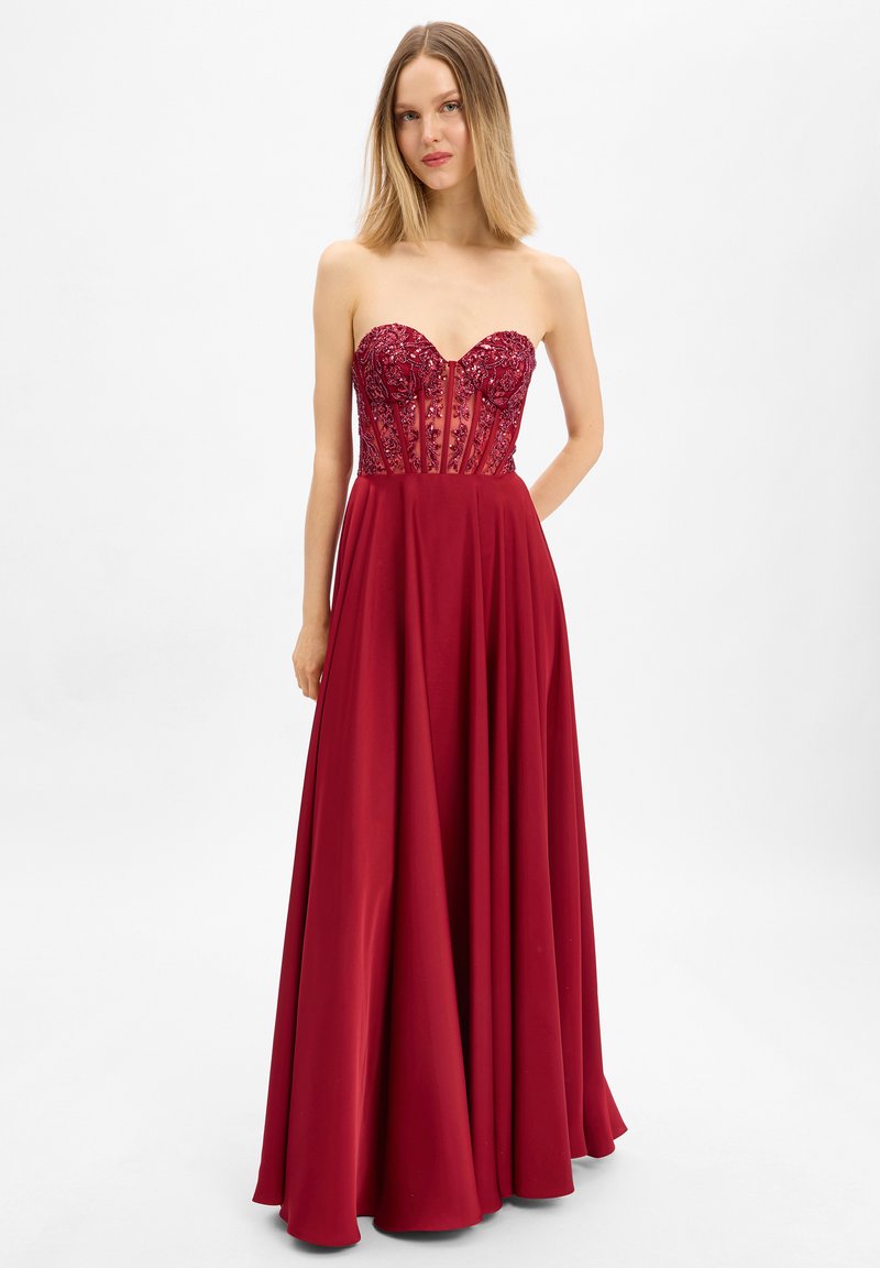 Femme portant une robe longue rouge foncé sans bretelles avec un corsage transparent orné et une jupe fluide, debout devant un fond blanc uni.