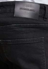 Mustadest denimist valmistatud teksad tekstuuriga pinnaga, millel on nahast silt "ZV PREMIUM" vööl ja standardne tagatasku disain.