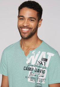 Camiseta verde claro con cuello en V, con texto impreso y elementos gráficos, que presenta acentos negros y una etiqueta de parche. Material suave y casual.