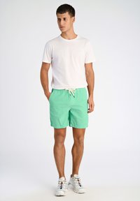 Costume da bagno verde a righe con vita regolabile, abbinato a una maglietta bianca e sneakers bianche. Design semplice e tessuto leggero.