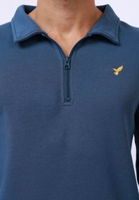 Námořnický modrý svetr s límečkem na zip, vyrobený z měkké tkaniny. Obsahuje malé logo žlutého ptáčka v oblasti levého prsou.