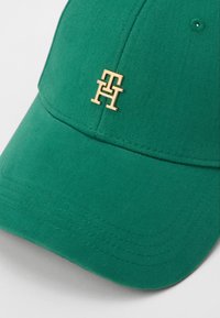 Căciulă din bumbac verde cu textură ribbed, având un logo auriu în față. Designul include un bor moale și detalii cusute.