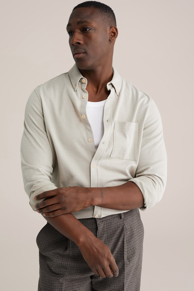 Šviesiai smėlio spalvos marškiniai su sagomis, krūtinės kišene, rankovėmis susuktais į viršų, derinami su tamsiomis languotomis kelnėmis. Audinys su tekstūra, neutralios spalvos.