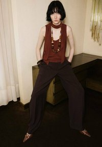 Top senza maniche marrone con dettaglio di sciarpa, abbinato a pantaloni larghi scuri e scarpe marroni a punta. Accessoriato con una collana di perline multicolori.