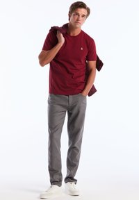 Bordeaux t-shirt met korte mouwen, grijze broek en witte sneakers. De t-shirt heeft een klein logo op de borst. Casual outfit.