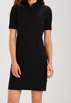 Mujer con un vestido tipo polo negro de manga corta, con cuello abotonado y un pequeño logo en el pecho izquierdo, sobre un fondo liso.