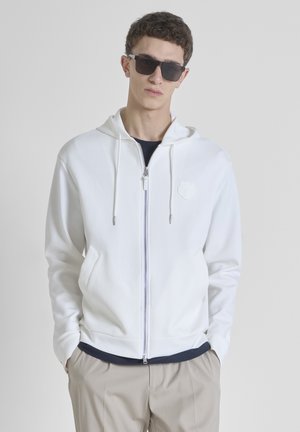 Haut à capuche blanc zippé, avec un logo texturé sur la poitrine, des poches avant et des finitions contrastées. Porté par-dessus une chemise sombre.