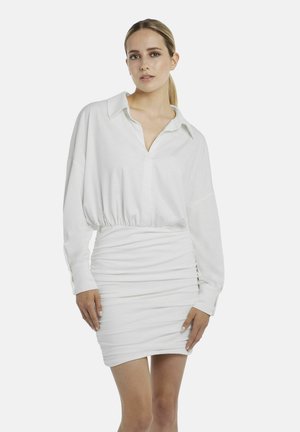 Vrouw met een lichte huid en blond haar naar achteren gebonden, gekleed in een witte top met lange mouwen en kraag en een gerimpelde witte mini-rok, staand tegen een effen achtergrond.