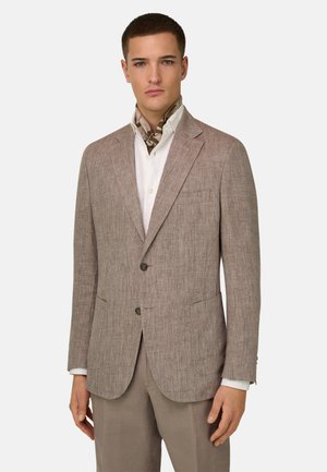 Mężczyzna ubrany w jasnobrązową, teksturowaną marynarkę na białej koszuli z wzorzystą apaszką na szyi oraz pasujące jasnobrązowe spodnie, stoi zwrócony przodem.