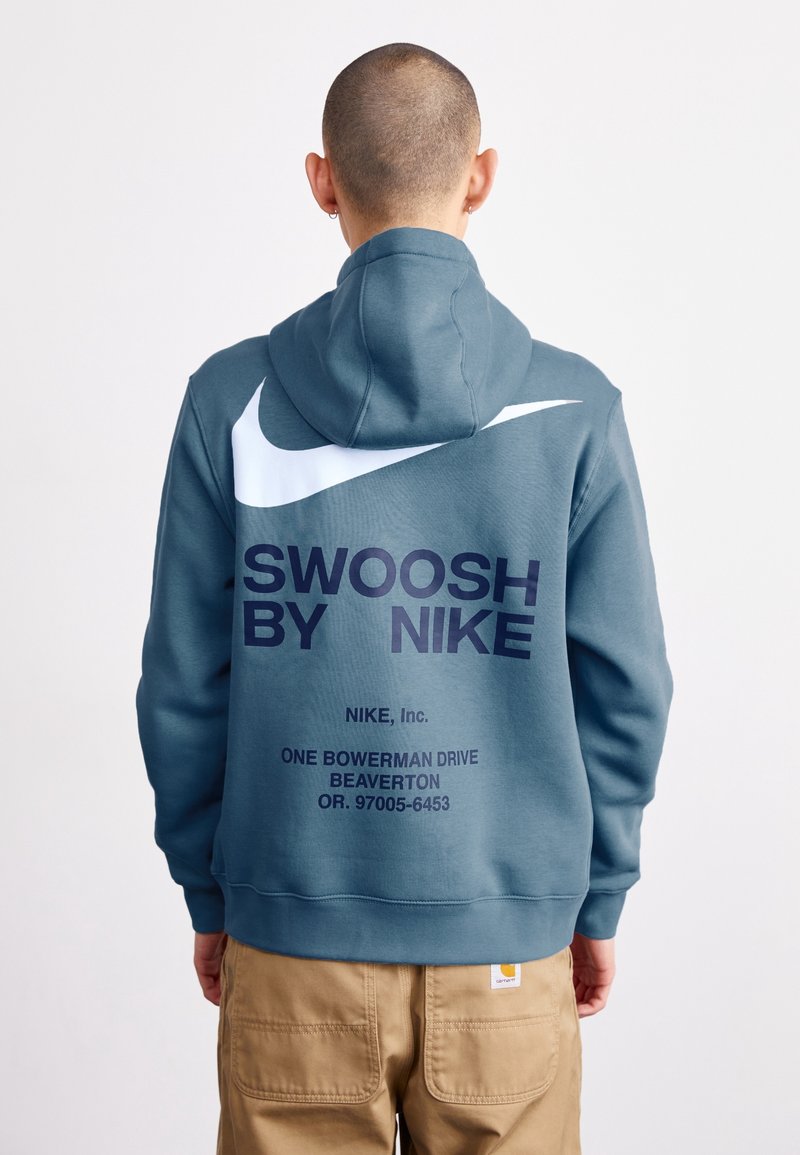 Huvtröja i teal-tyg har en stor vit Nike swoosh och fet text på ryggen, med ribbade mudd och söm.