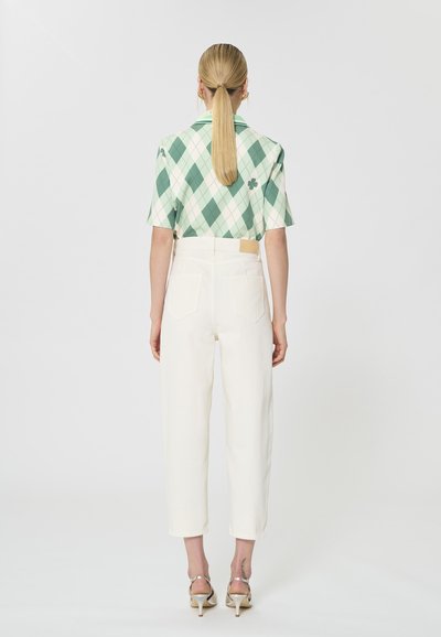 Femme avec queue de cheval blonde portant une chemise à manches courtes à losanges verts et blancs, un pantalon blanc taille haute et des talons beiges, debout de dos.