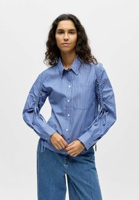 Chemise boutonnée rayée bleu et blanc avec manches froncées et détails à cordon, dotée d'une poche sur la poitrine gauche, en coton.