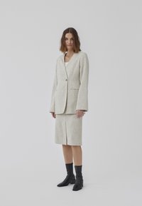Costum din in de nuanță bej deschis, cu sacou cu rever clasic, închidere cu un singur nasture și fustă până la genunchi cu fendue frontală, asortată cu botine negre.