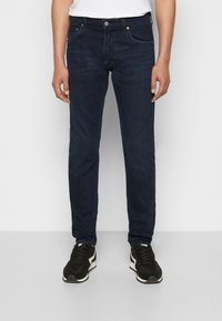 Marinblå denimjeans med rak passform, fem fickor och orange sömmar, kombinerat med svarta sneakers med vita detaljer.