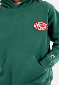 Sudadera verde de tela suave con un bolsillo frontal y un parche ovalado rojo que dice "Sweet Pants". Incluye un detalle redondo en el puño.