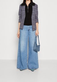 Blazer à carreaux avec un col noir, haut texturé noir, jean bleu à jambes larges et petit sac à main bleu, avec fermeture éclair et détail en forme d'anneau.