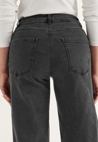 Grå denimjeans med hög midja, fyra fickor och en lätt slitna textur. Bakifrån framhävs fickornas placering och sömmarna.