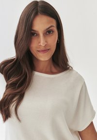 Bílý top s krátkým rukávem vyrobený z hladké, lehké tkaniny, s kulatým výstřihem a uvolněným střihem, s jemnou texturou.