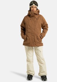 Braune isolierte Jacke mit Reißverschlusstaschen, getragen über beigen Schneehosen. Das Modell trägt schwarze Stiefel und eine Schutzbrille sowie eine gestrickte Mütze.