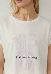 T-shirt en coton blanc avec un contour violet clair de tulipes dans un vase et le texte "RUE DES FLEURS" en bleu foncé. Manches courtes.