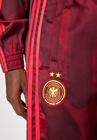Maroon sportska jakna sa crvenim prugama, rebrastim manžetama i okruglim izvezenim logotipom s orlom i zvijezdama.