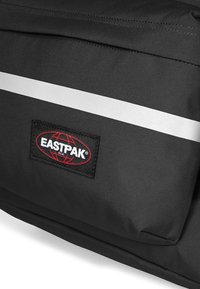 Mochila de tela negra con una superficie texturizada, que presenta una franja reflectante blanca y un parche de logo de Eastpak en rojo y negro.