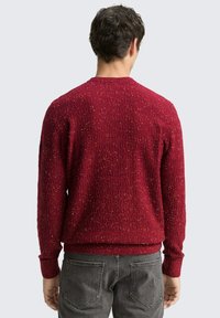 Pull en laine rouge avec un motif texturé, col rond et manches longues. Présente des poignets et un ourlet en côtes. Porté avec un jean gris.