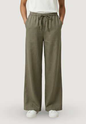 Persona che indossa pantaloni a gamba larga verde oliva con coulisse, con le mani nelle tasche e sneakers bianche su uno sfondo semplice.