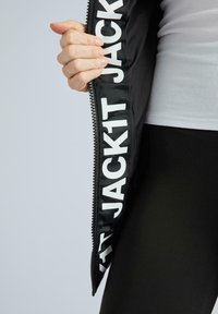 Juodas švarkas su dideliu baltu užrašu "JACK1T" vidinėje pamušalo pusėje. Matyti užtrauktuko detalė, dėvimas su baltu tekstūruotu marškinėliu ir juodomis legging'omis.