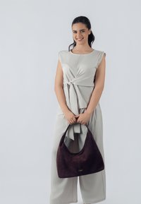 Bolso hobo en ante púrpura oscuro con forma curva. El modelo lleva una blusa sin mangas de color gris claro con detalle anudado y pantalones anchos.