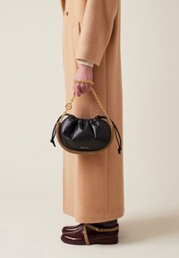 Schwarze Lederhandtasche mit gerafftem Oberteil, goldener Kettenhenkel und strukturiertem goldenen Besatz. Getragen von einer Person in einem langen beigen Mantel und kastanienbraunen Stiefeln.