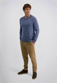 Hellblauer Strickpullover mit geripptem Ausschnitt, beige Cordhosen und braunen Wildledersneakern. Mann steht mit den Händen in den Taschen.