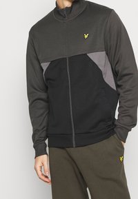 Chaqueta con cremallera en gris y negro, con diseño a bloques de color, puños acanalados, cuello alto y pequeño logo amarillo en la parte superior del pecho.