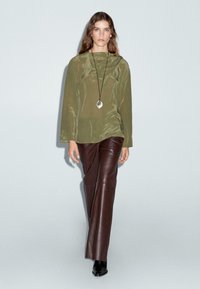Haut à manches longues en velours vert olive, associé à un pantalon en cuir marron évasé et des bottines noires. Collier avec pendentif en argent comme accent.