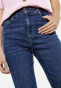 Kõrge vöökoht, tummiselt tumesinised teksapüksid, millel on kitsas lõige, tõmblukuga esiosa, viis taskut ja peened õmbluse detailid.