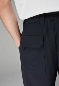 Navy jogger-housut, joissa on joustava vyötärö, sivutasku läpällä ja sileä pinta. Materiaali vaikuttaa kestävältä ja kevyeltä.