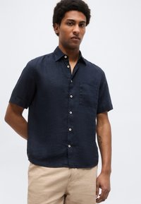 Άνδρας με σγουρά μαλλιά, ντυμένος με μπλε κοντομάνικο πουκάμισο με κουμπιά και μπεζ παντελόνι, στέκεται με το ένα χέρι πίσω από την πλάτη του.