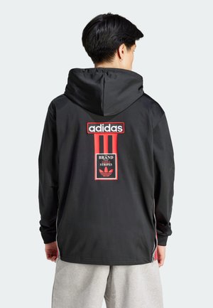 Osoba nosząca czarną bluzę z kapturem Adidas z czerwono-białym logo i napisem „The Brand with the Three Stripes” na plecach oraz jasnoszare spodnie.