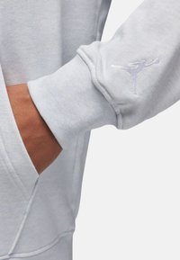 Lys grå sweatshirtdetalje med en ribbet manchet og et broderet logo af en figur i luften, der fremviser et blødt, struktureret stof.