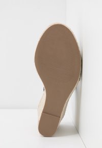 Semelle d'une sandale beige à talon compensé avec une semelle en caoutchouc texturée, positionnée contre un angle de mur et de sol blancs.
