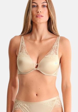 Femme portant un soutien-gorge en dentelle beige et une culotte assortie, tournée vers l'avant sur un fond gris clair.