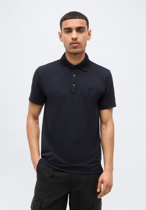 Jeune homme portant un polo noir à manches courtes et un pantalon cargo noir, debout devant un fond gris clair uni.